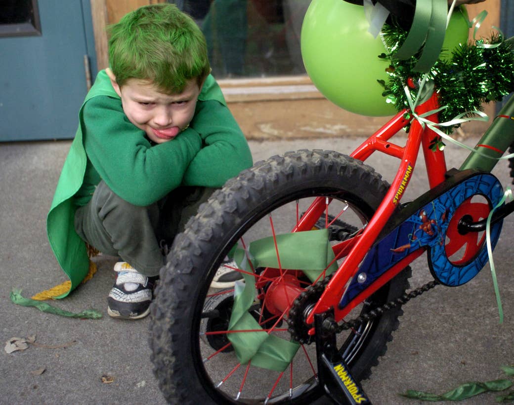a kid with green hair and wearing green clothes sticks his tongue out and squats next to a bike with green ribbons and a balloon 