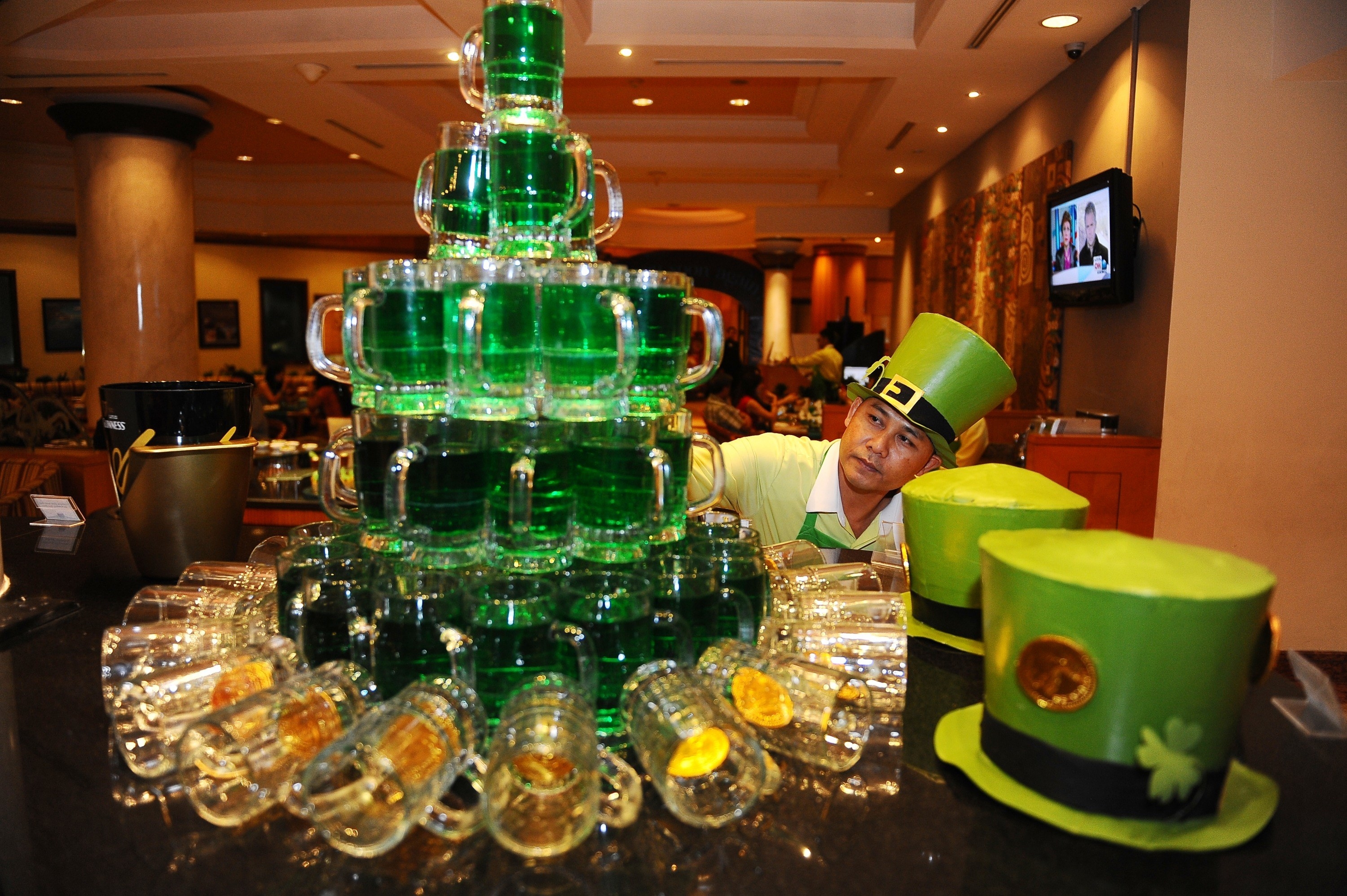 a green beer tower with two hats on the right and a man adjusting it in a st patrick&#x27;s day hat on the right