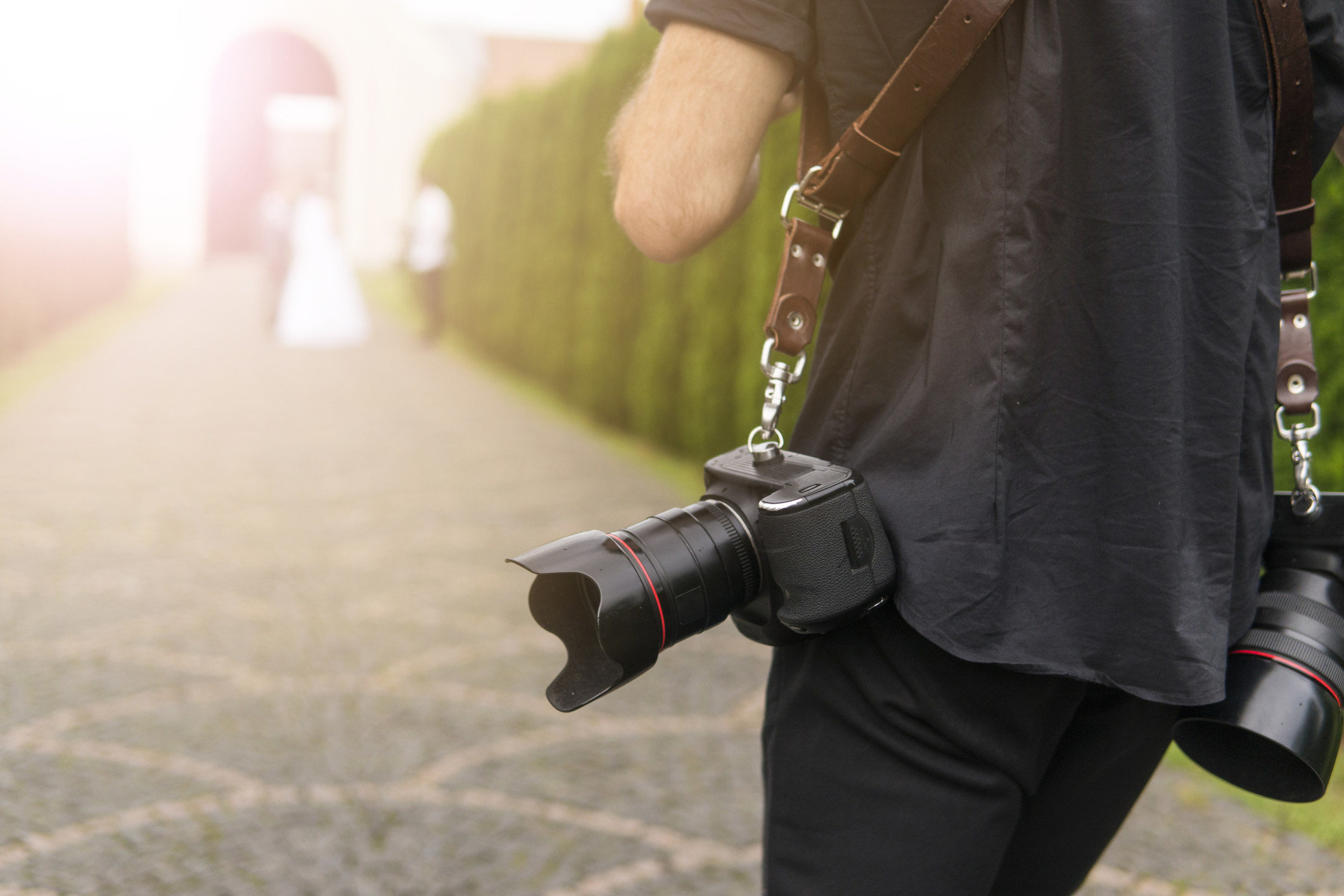 A wedding photographer with two cameras follows a bride and groom down a path