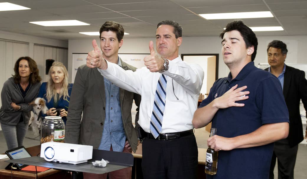 A man standing between two other men in an office gives two thumbs up