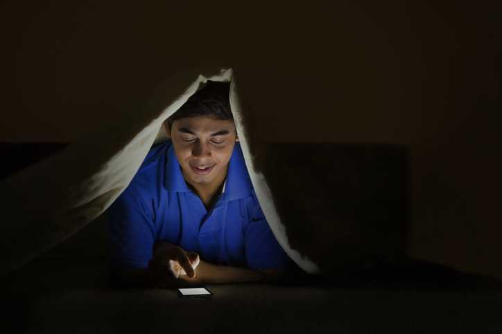 A guy sitting up under the covers in bed in the dark and looking at his phone