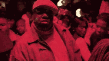 Biggie wearing a newsboy cap and nodding along to music at a club full of people
