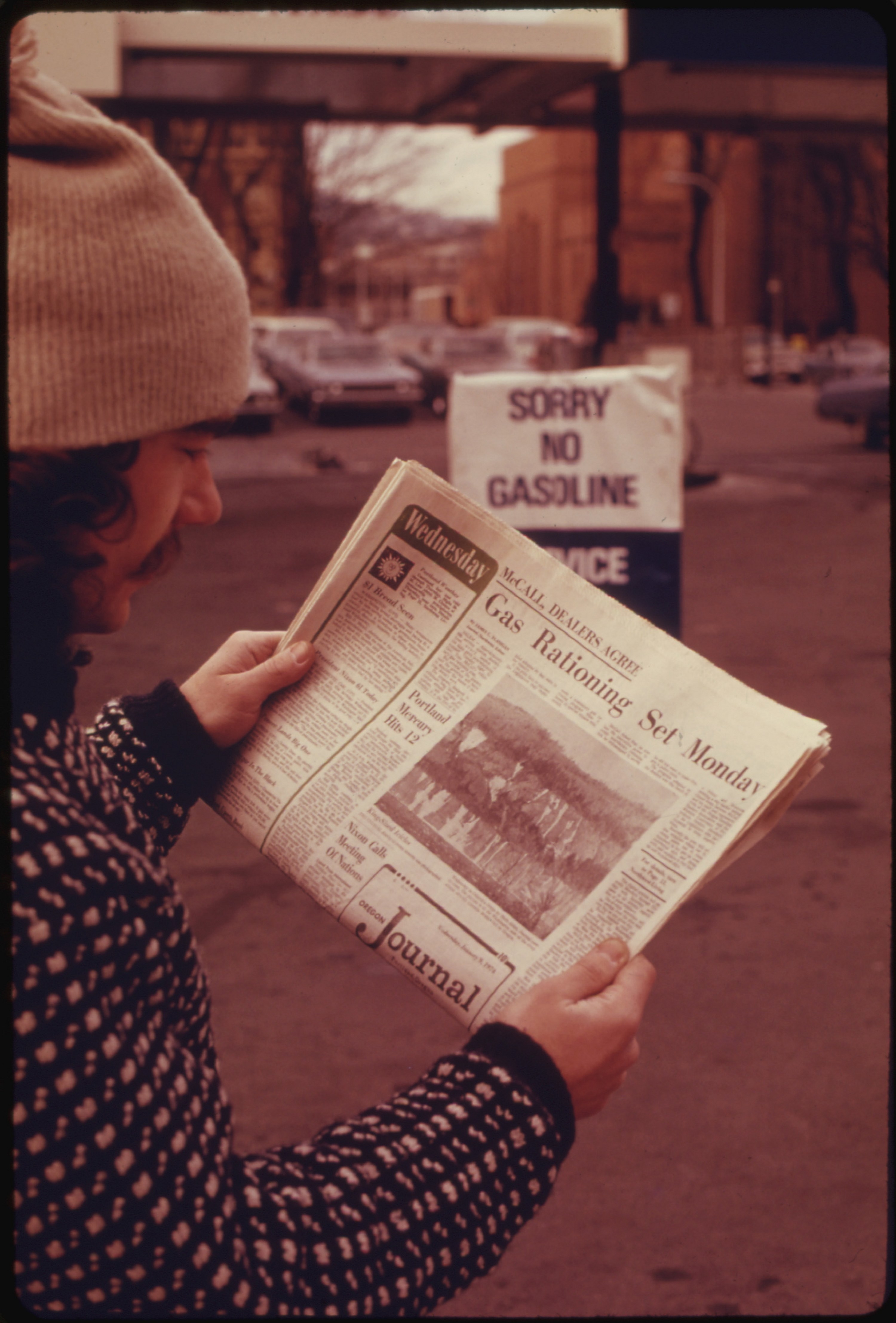 A person with a mustache wearing a beanie reads a newspaper with the headline &quot;Gas Rationing Set Monday&quot;