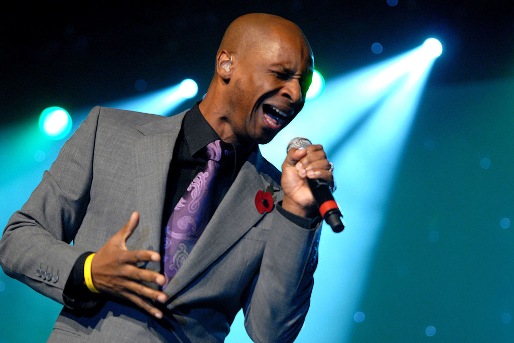 Andy Abraham in a grey suit, black shirt, and purple tie. He is holding a microphone in one hand with his eyes closed