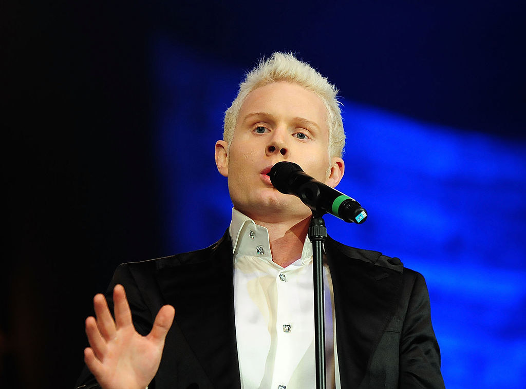 Rhydian Roberts in a black blazer and white shirt undone, singing. A mic stands in front of him and he has his right hand raised in front of him