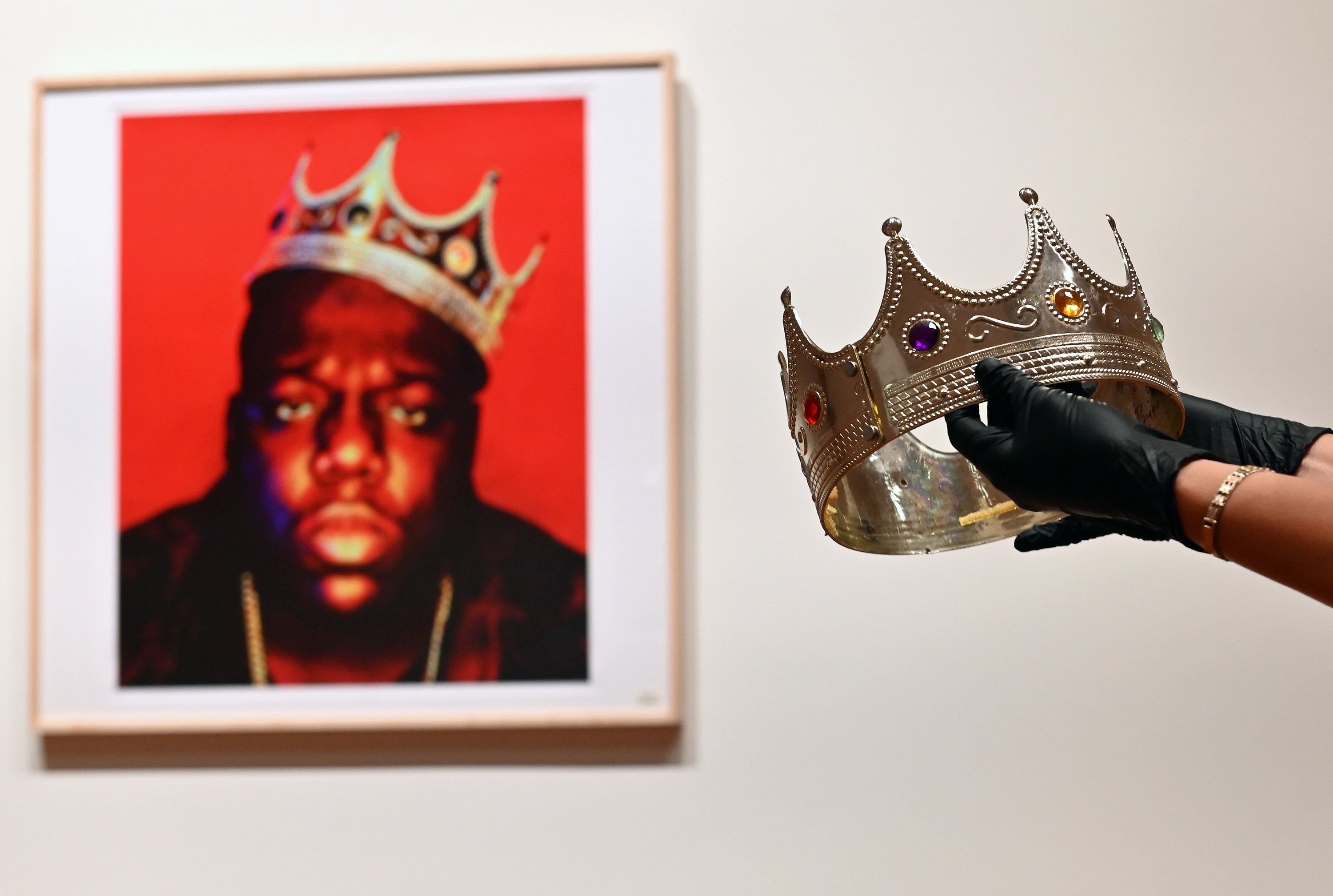 gloved hands hold out a crown toward a photo of Biggie wearing a crown