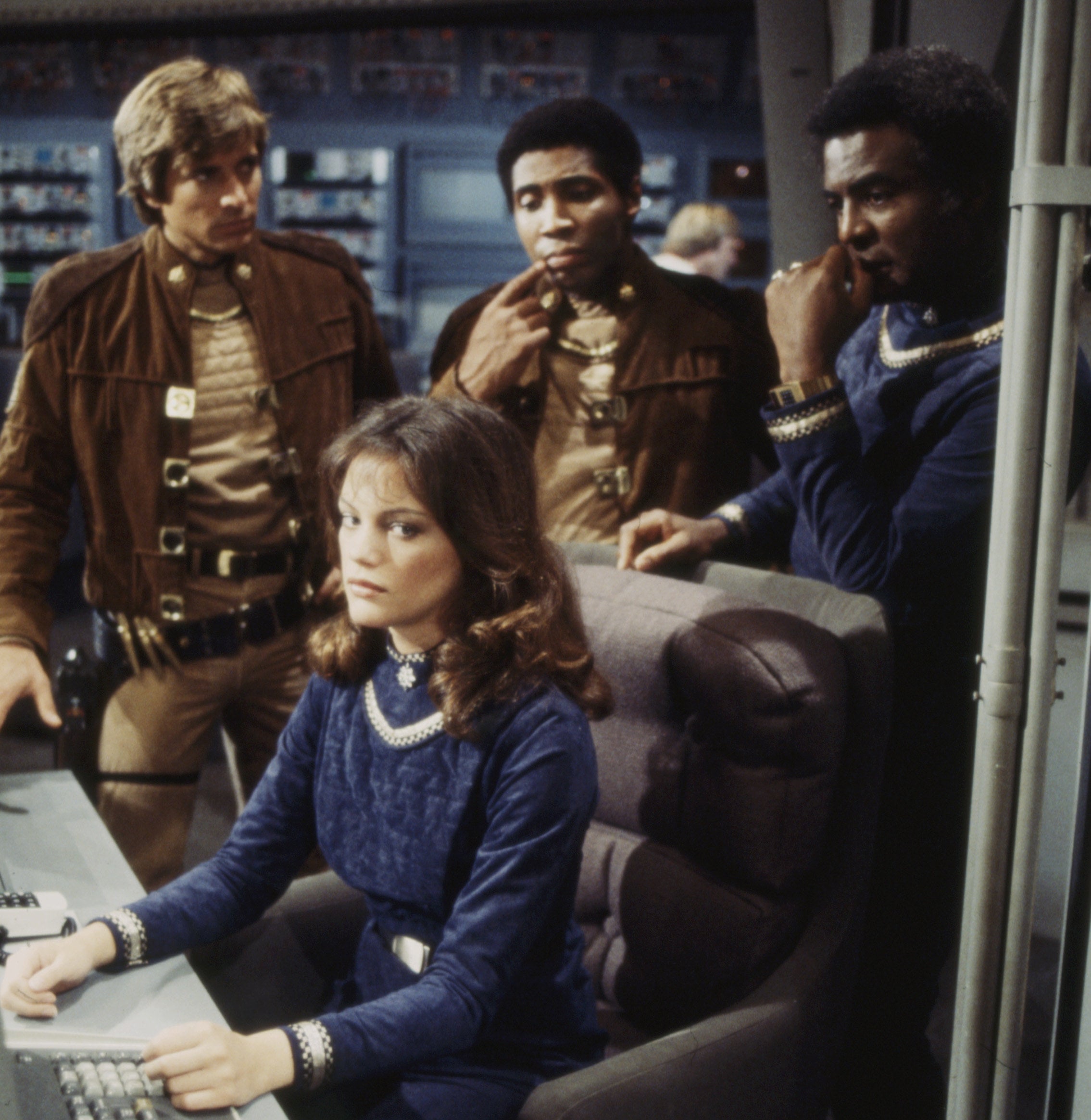 The original Battlestar Galactica cast standing over a woman sitting at a table with what looks like a calculator