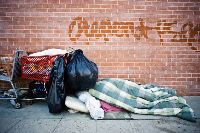 someone sleeps under blankets and next to a shopping cart filled with items on a street
