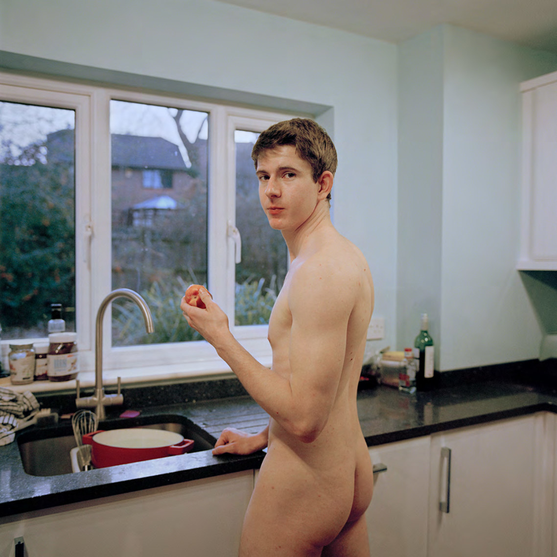 A naked man standing in front of a window at a kitchen sink and holding a fruit looks at the camera