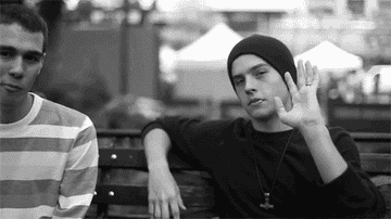 Dylan Sprouse waving at the camera, wearing a beanie and chewing on a toothpick