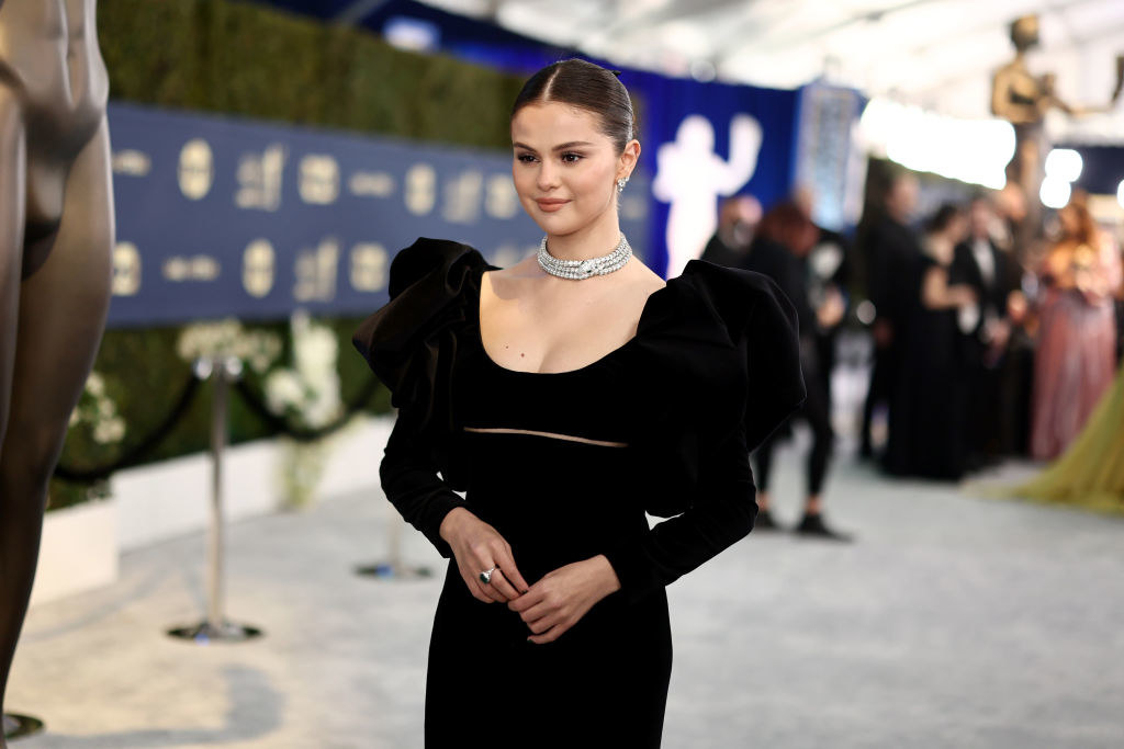 Selena sports a sleek bun, a chunky diamond choker necklace, and a long sleeved dress with large, puffed out shoulders