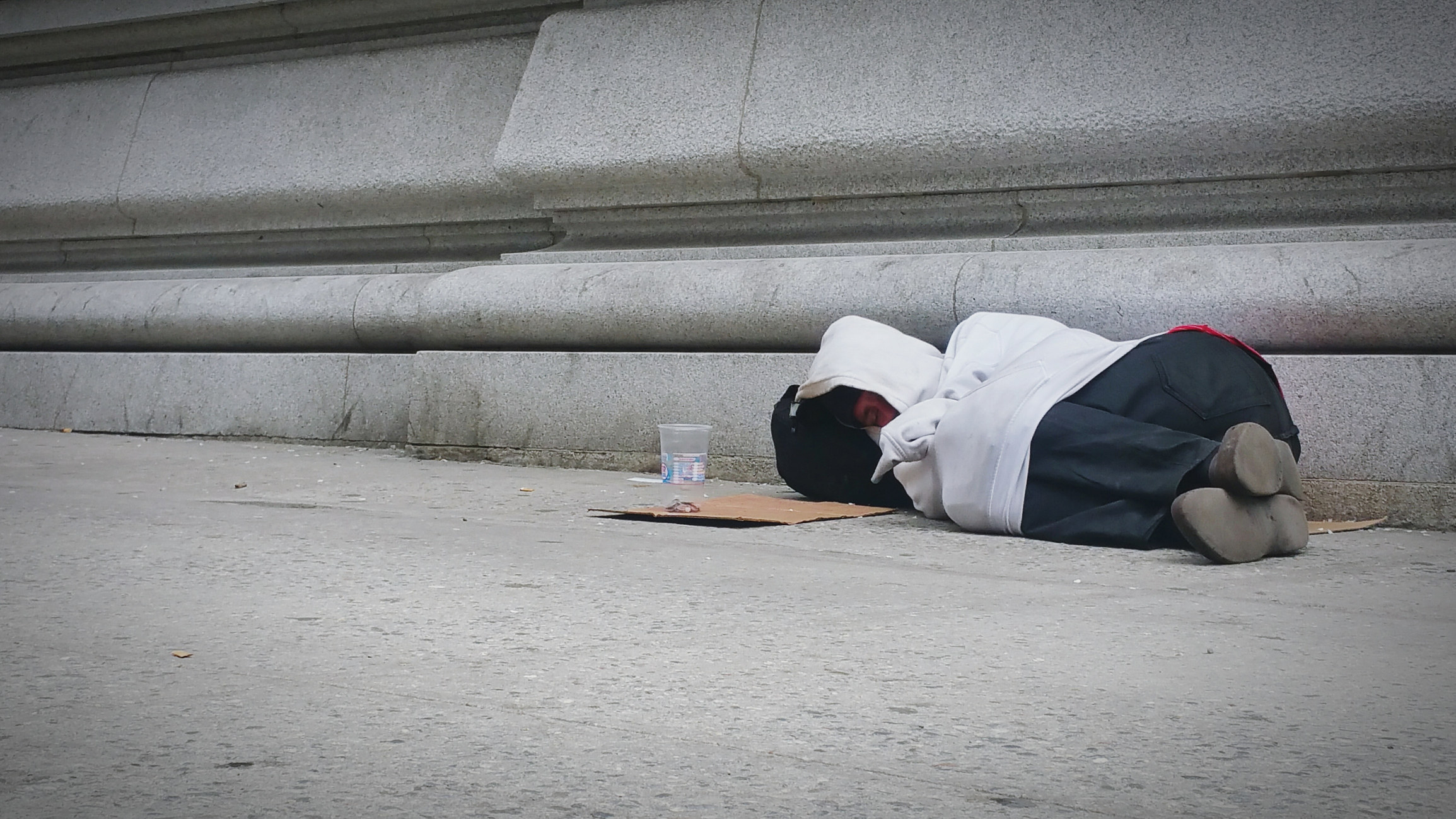 someone sleeps outside on the street with cardboard and a cup next to them