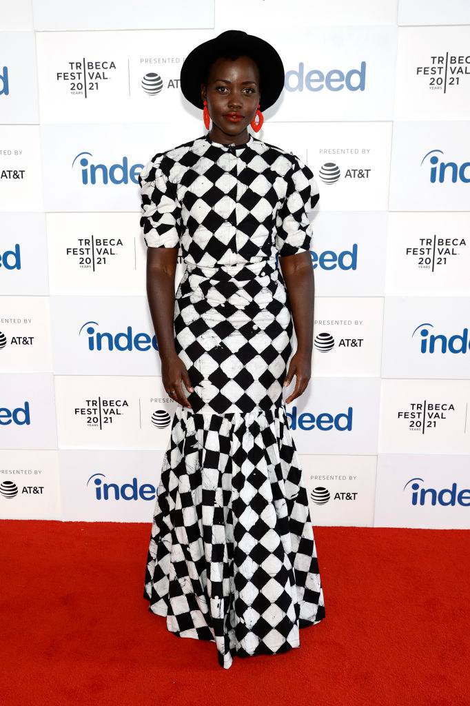 Lupita wears a bowler hat, chunky neon hoop earrings, and a '50s-inspired checkered mermaid gown with cap sleeves