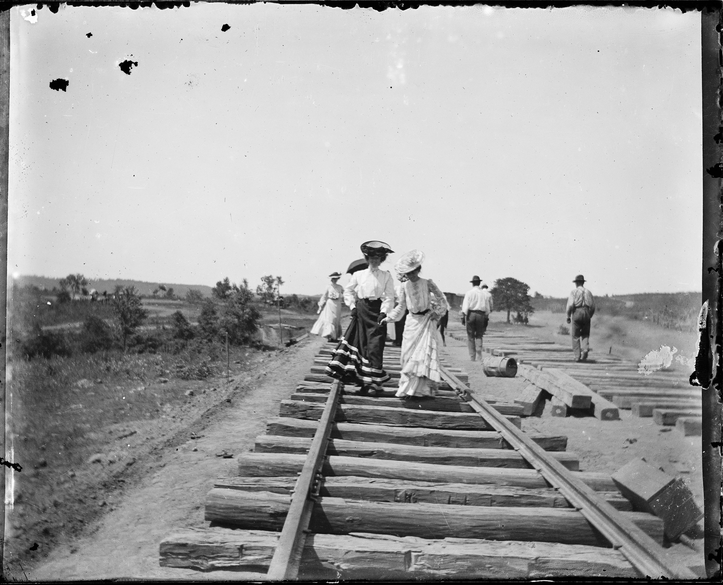 Women in turn-of-the-century hats and long dresses, with men wearing overalls and hats behind them, walk along train tracks under construction in Oklahoma in 1902
