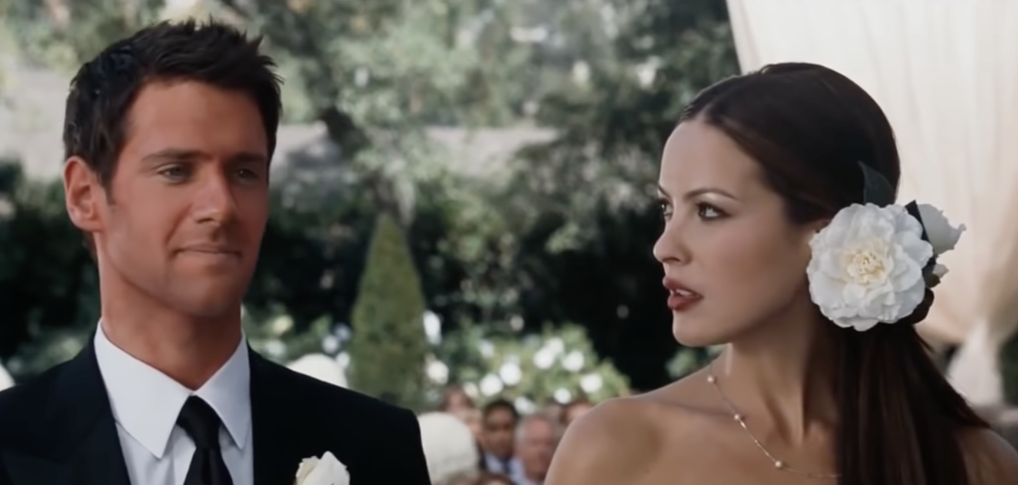 Two people at a formal outdoor event. The woman wears a floral hair accessory and strapless dress; the man is in a suit