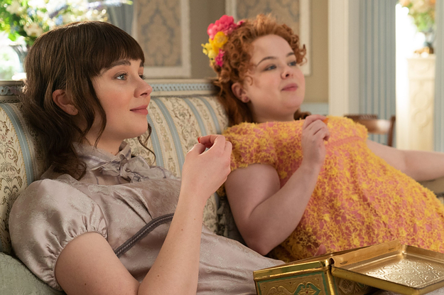 Eloise Bridgerton and Penelope Featherington sit side by side on a couch eating chocolates from a tin box