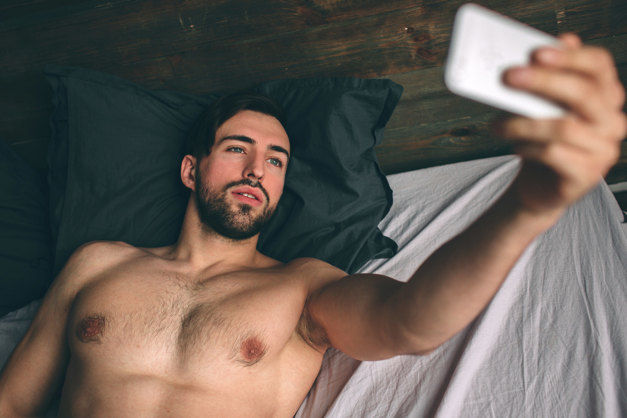 Naked, bearded, dark-haired handsome man shirtless in white bed taking a selfie