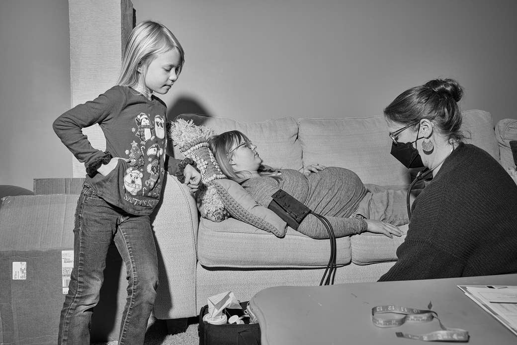 A pregnant woman lies on the couch with a blood pressure cuff as a midwife attends to her and her daughter looks on 