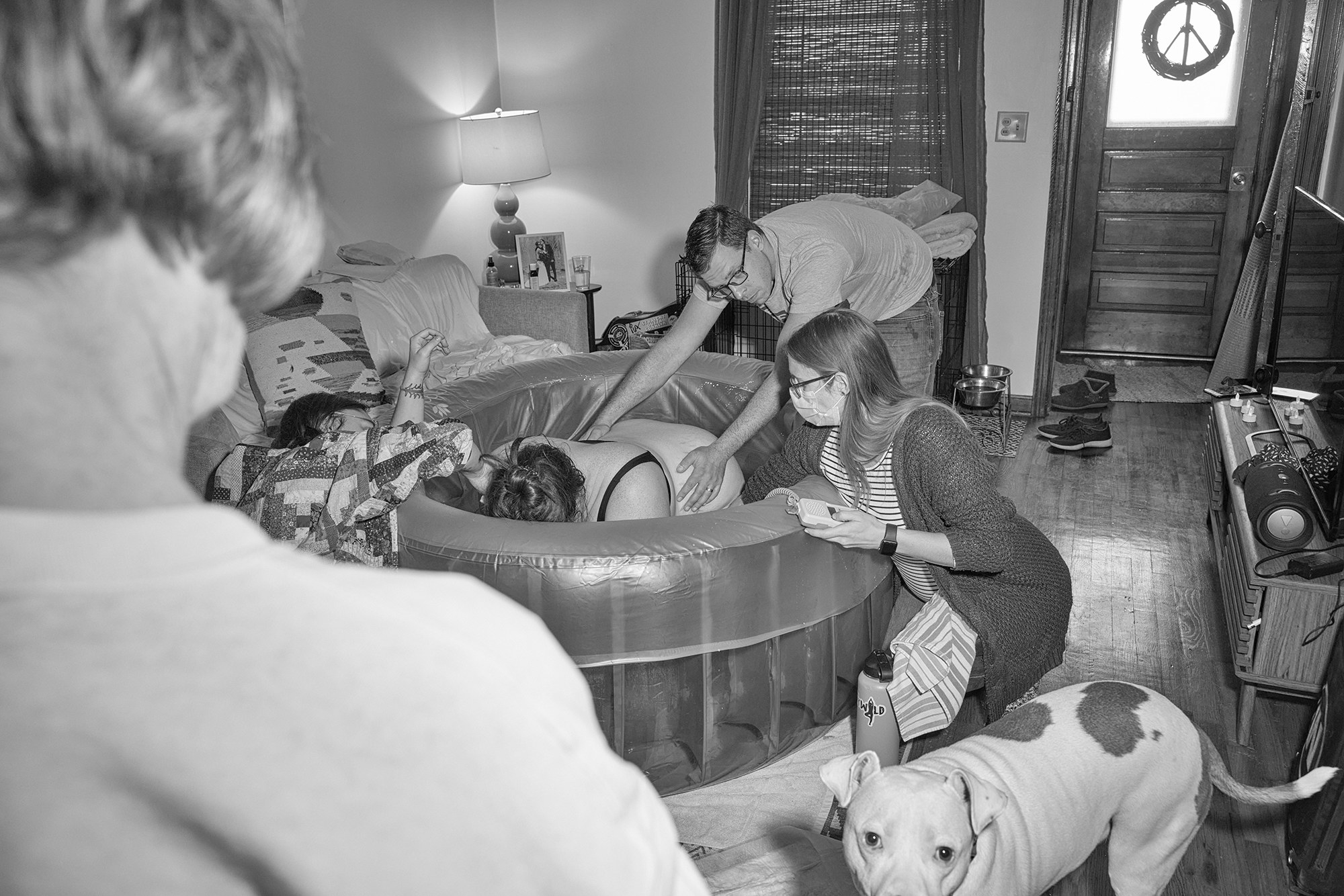 A woman on all fours in a birth tub while midwives look on and her dog walks around