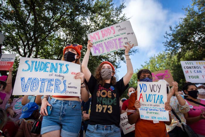 People wearing masks hold up various signs, including "My Body My Choice My Vote My Voice" and "Students of Today Voters of Tomorrow"