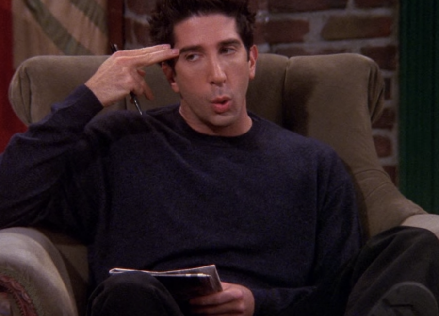 Ross Geller wears a dark colored sweater while holding two fingers to his temple