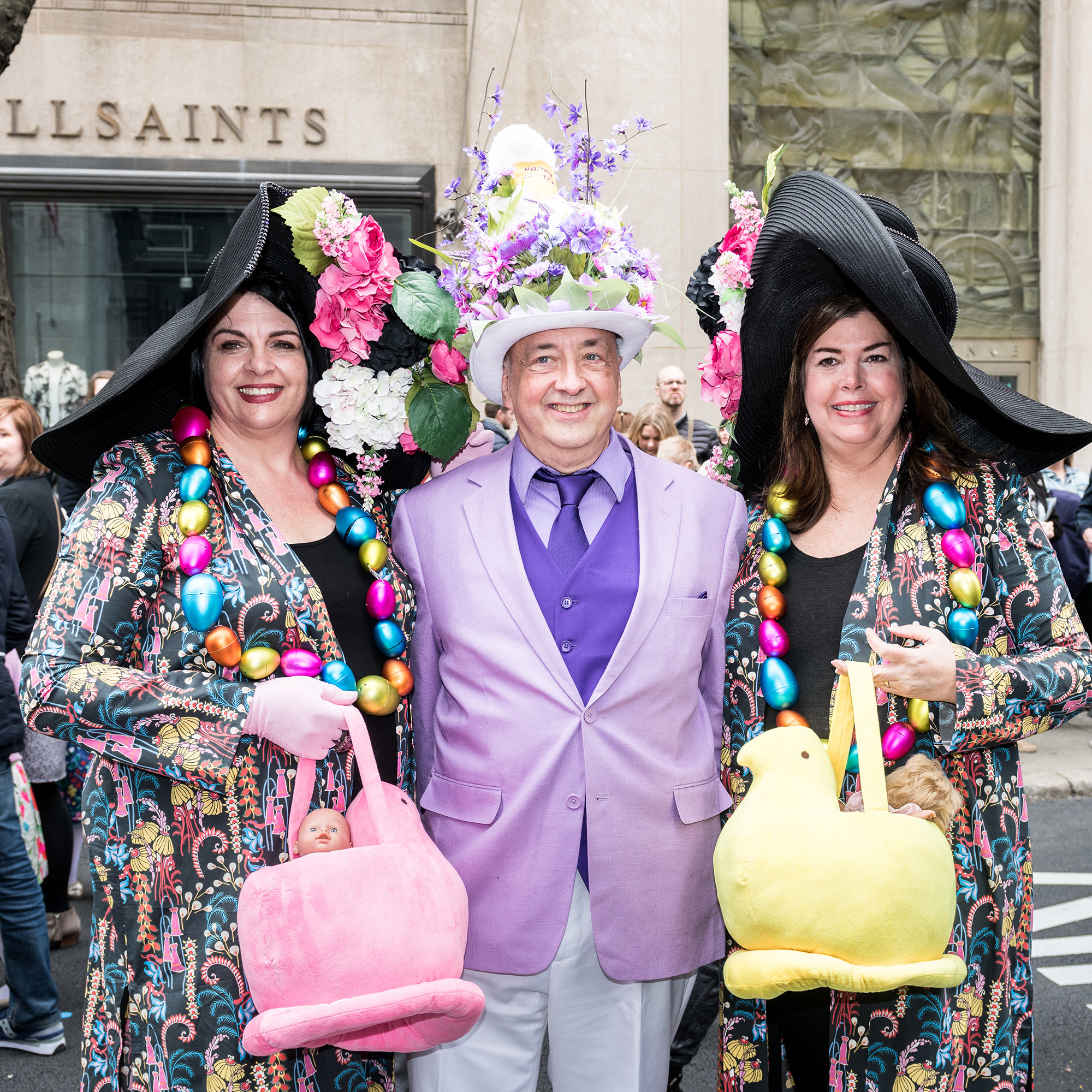 Two women in black hats and necklaces made of Easter eggs carrying baskets that look like peeps pose with a man in a purple suit 
