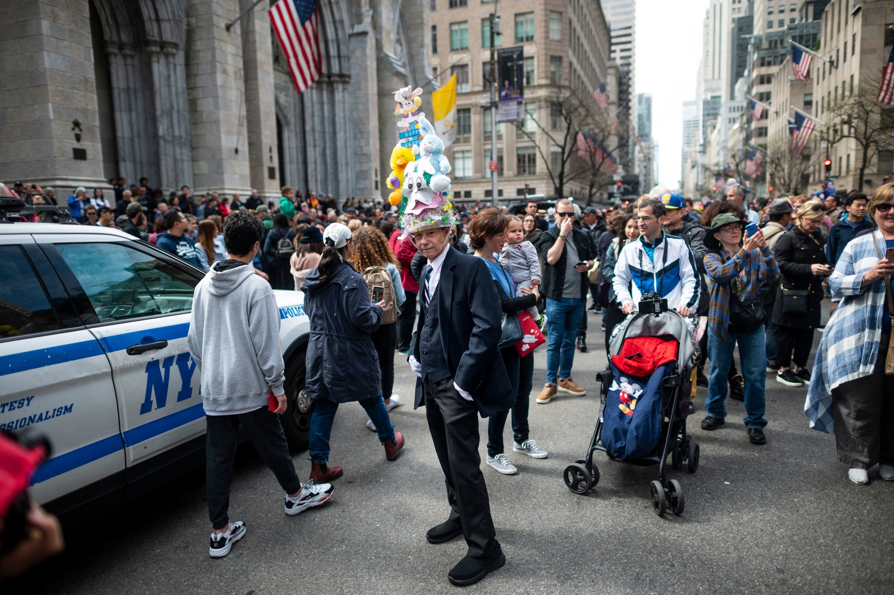 A man in a very high hat decorated with stuffed animals stands by an NYPD car as many people walk by