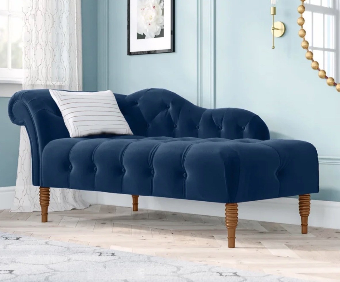 a tufted teal velvet chaise lounge with a scalloped art and wooden legs and a decorative throw pillow on top