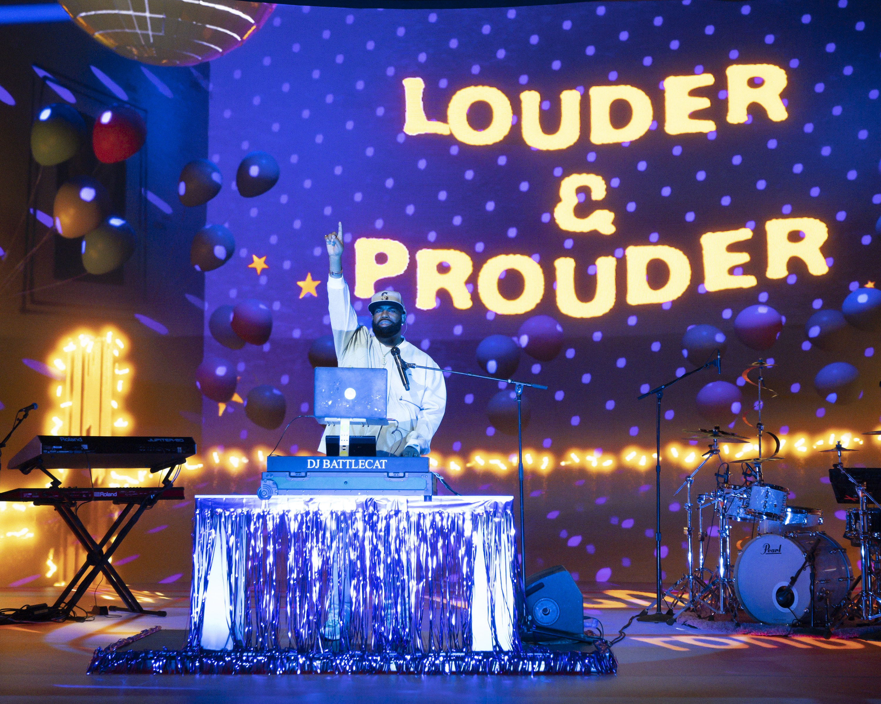 DJ Battlecat raises one finger in the air while spinning at turntable on stage in front of instruments, balloons, and a project light that reads &quot;Louder &amp; Prouder.&quot;
