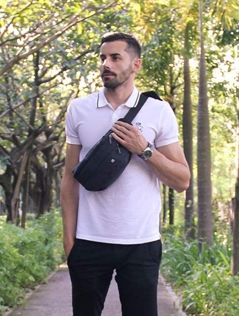 Someone standing against a forest background, posing with one hand on the strap of the belt bag that&#x27;s slung across their chest