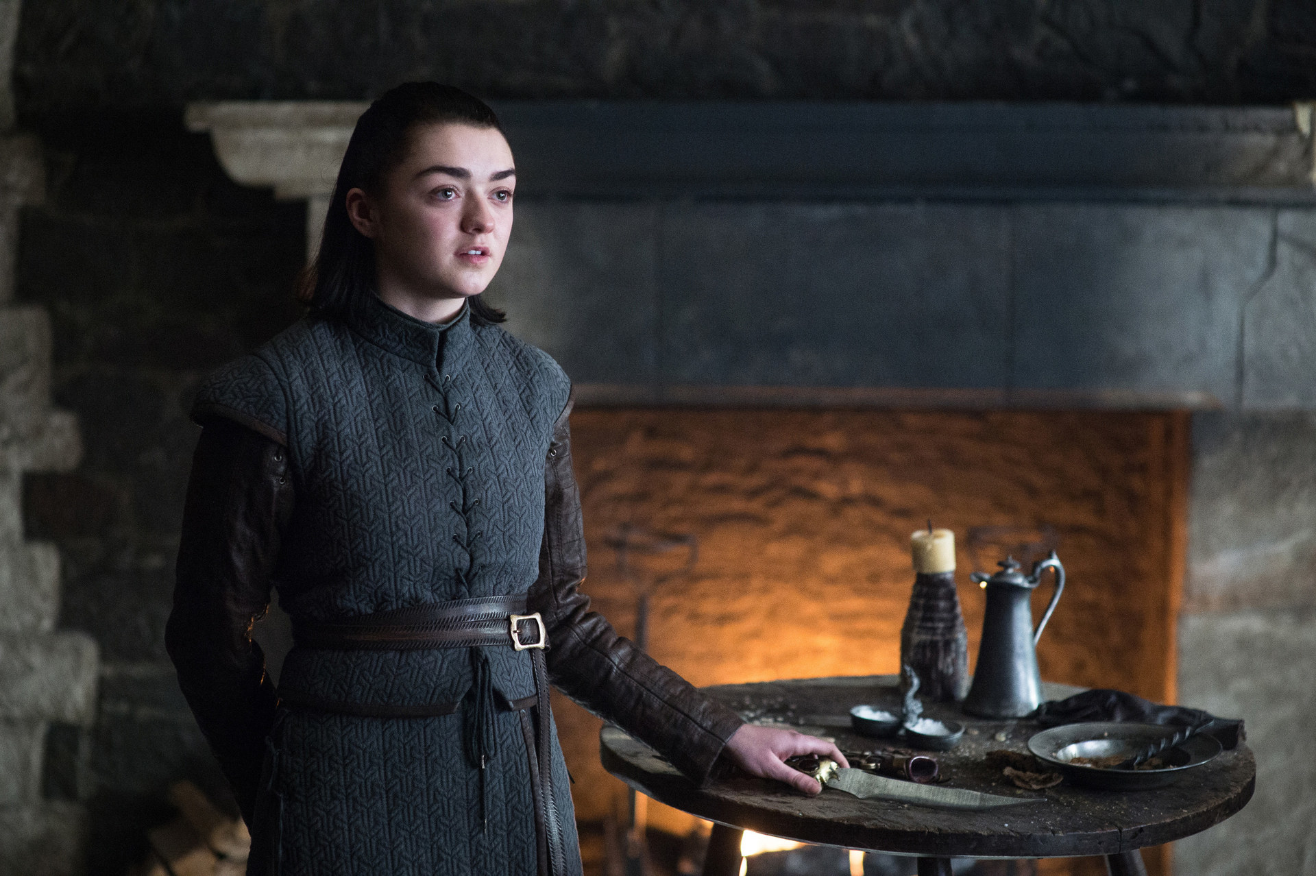 Arya stands near a table with a fire in the background