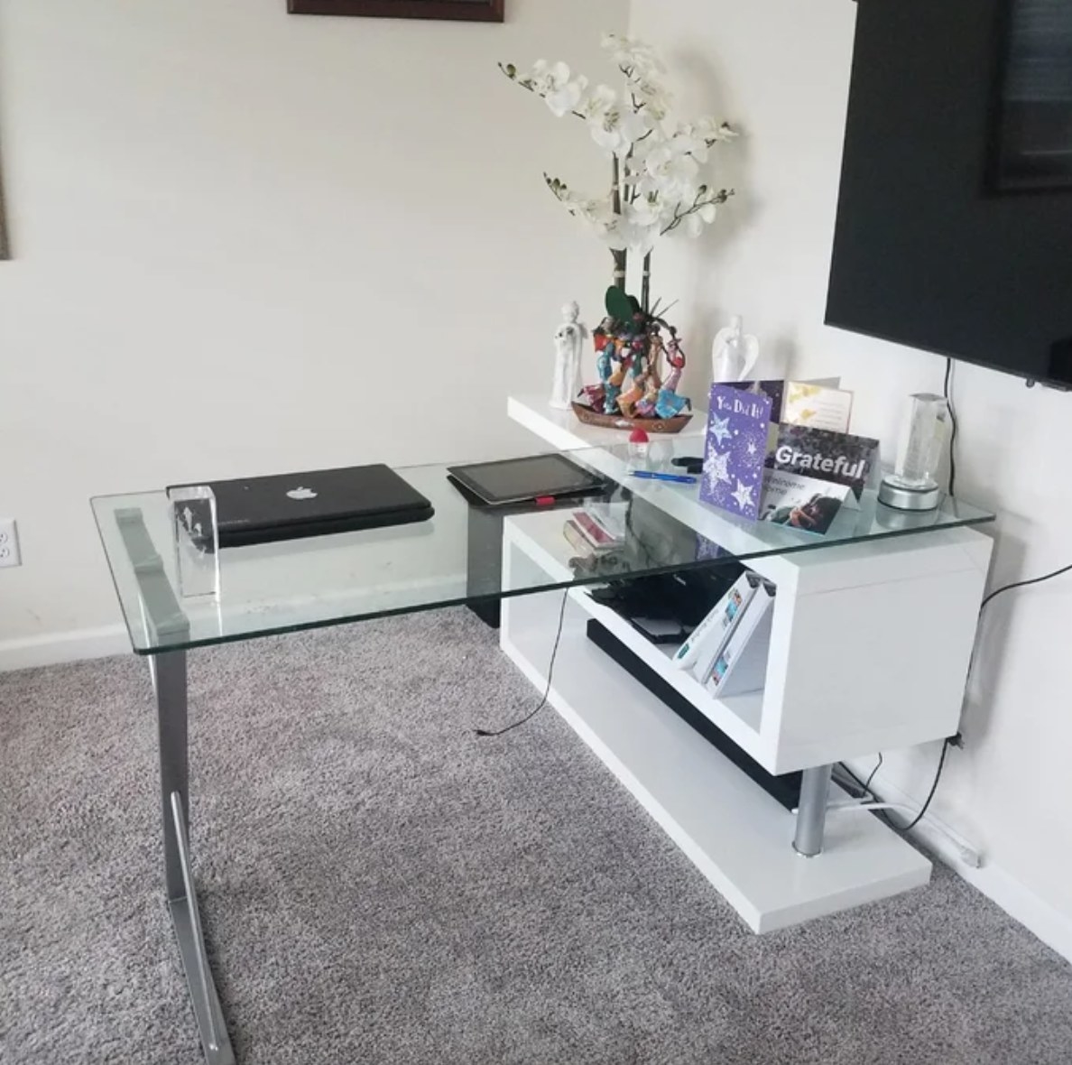 a white and clear L-shaped desk with a laptop, tablet, cards, and a flower arrangement on top