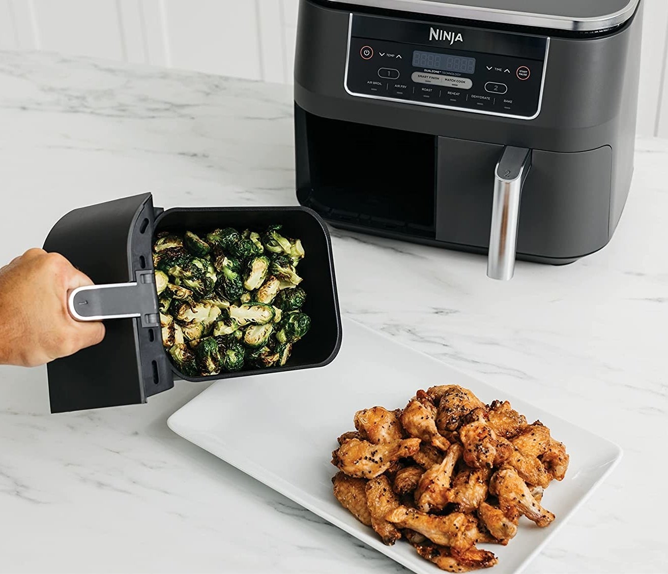Someone plating up some vegetables from one of the fryer baskets onto a plate of chicken wings, with the Ninja fryer behind it