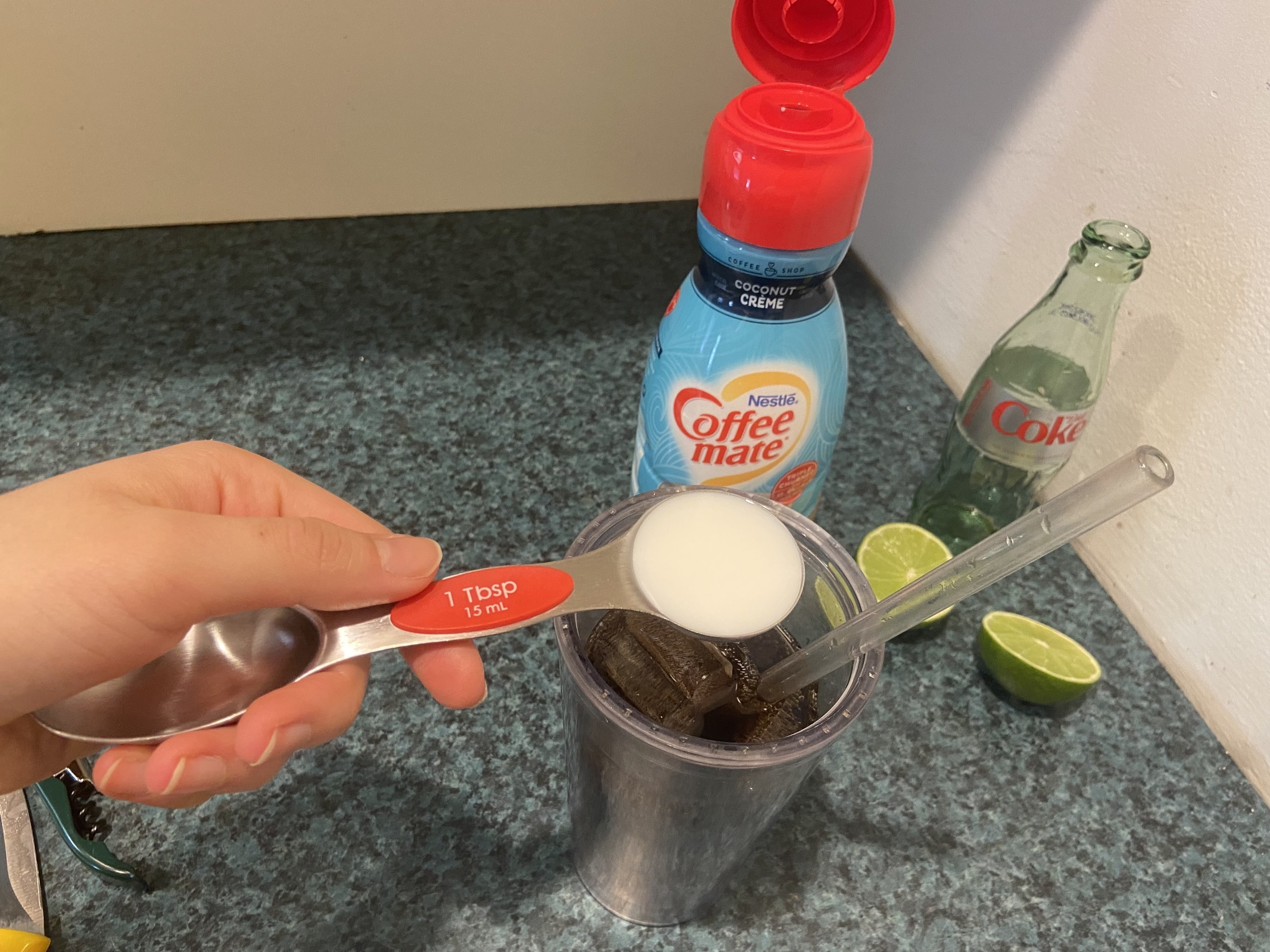 The author&#x27;s hand holding a measuring spoon of coffee creamer over a cup of Diet Coke