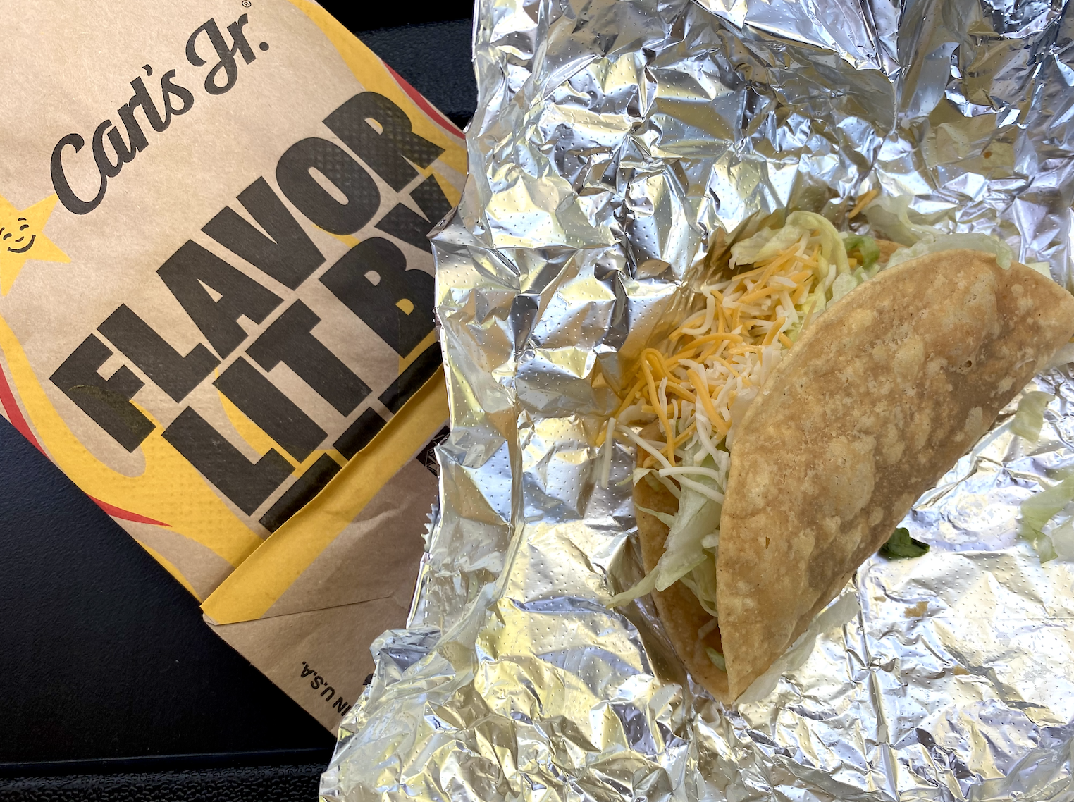 A hard shell taco with lettuce and cheese, on foil and on a Carl&#x27;s Jr&#x27;s paper bag