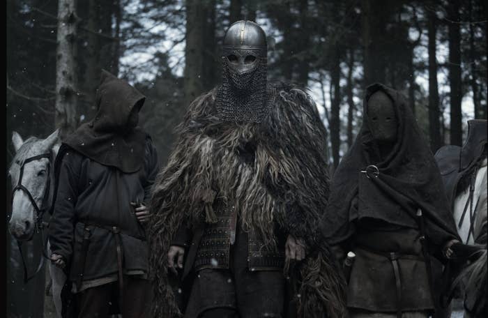 A man wearing a face covering helmet, and two masked and hooded men stand behind him