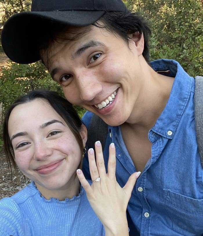 A young couple takes a selfie, the woman holding up an engagement ring