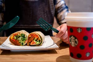 A burrito cut in half as two hands hold a knife and fork over it next to a take-out cup of coffee