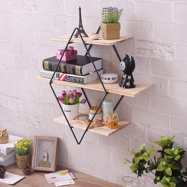 An image of an iron and wooden wall shelf with plants, books, and candles on the shelves