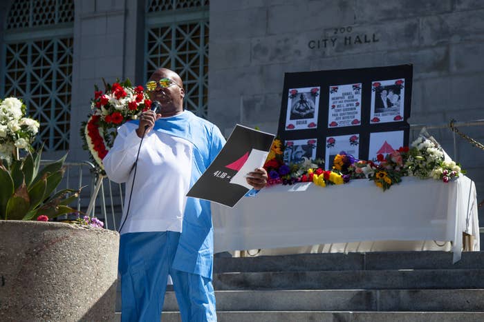 A man speaks into a microphone at the bottom of steps with a memorial with posters and flowers at the top