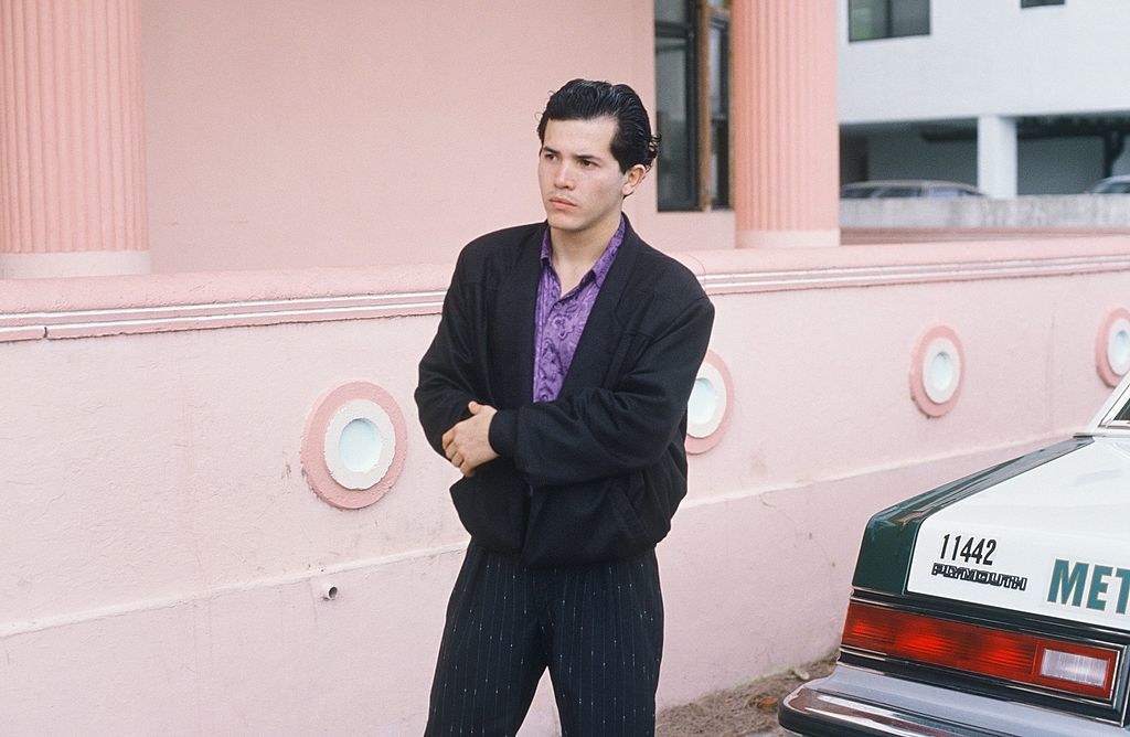 John Leguizamo standing with his arms crossed by a car