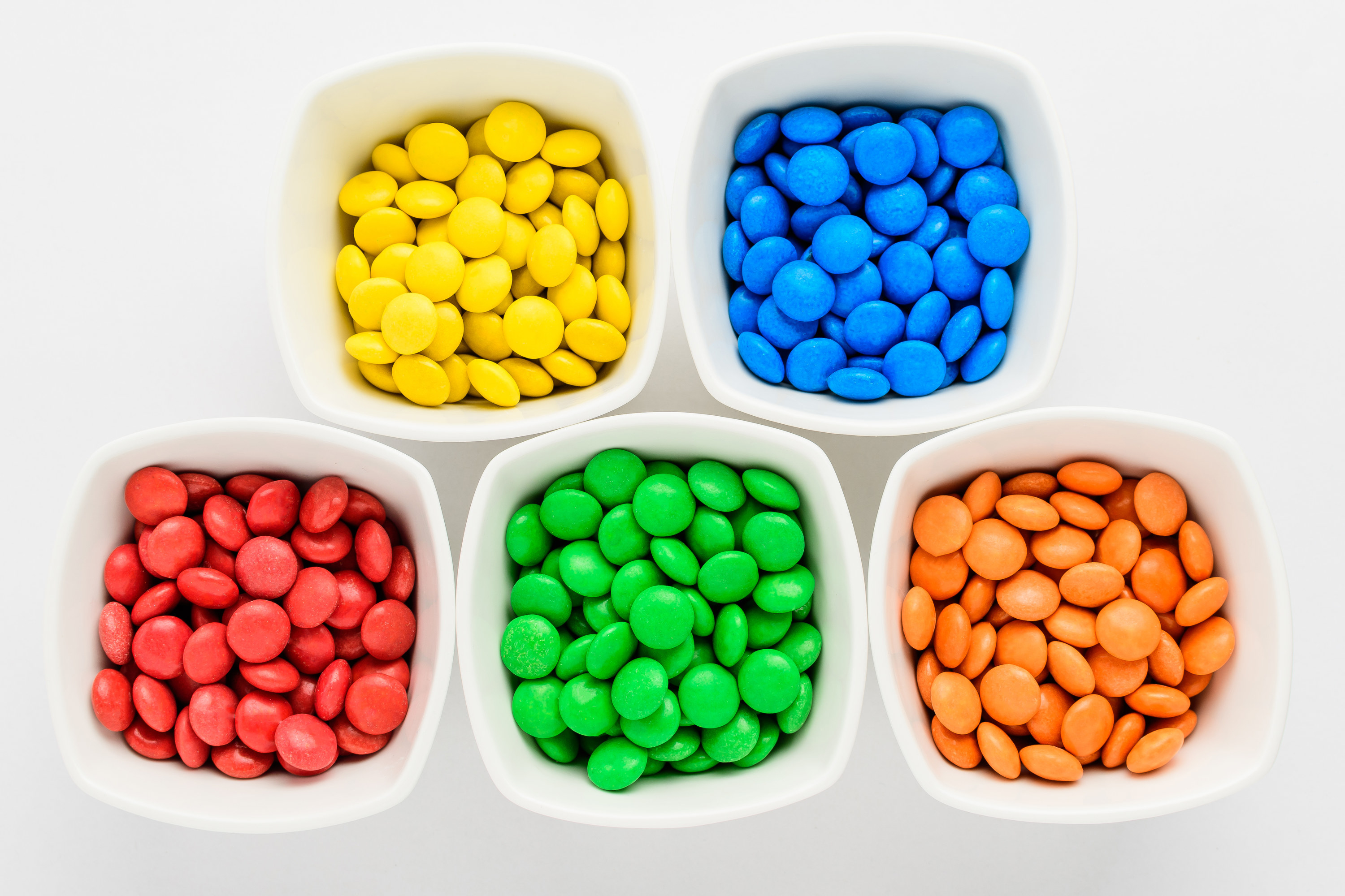 Five squared bowls with small red, blue, green and orange coated chocolate candies similar to m&amp;ms in a squared bowl isolated on white background, top view