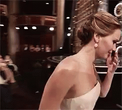 GIF of Jennifer Lawrence wearing a long white dress; she approaches the stairs to the stage and trips on the second one, falling to her hands and knees