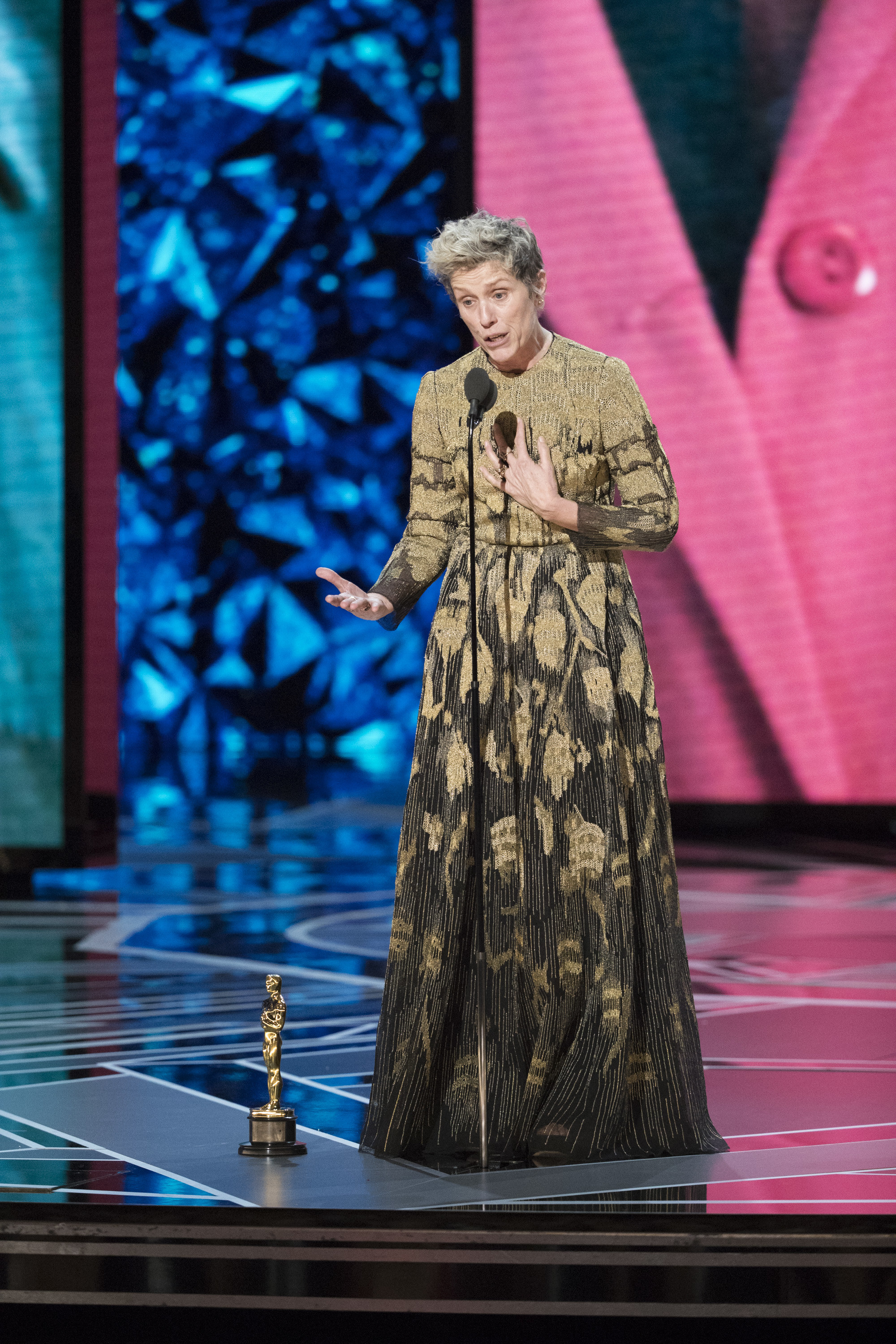Frances McDormand wears a long black and gold dress and speaks into a microphone onstage, her Oscar statue on the floor in front of her