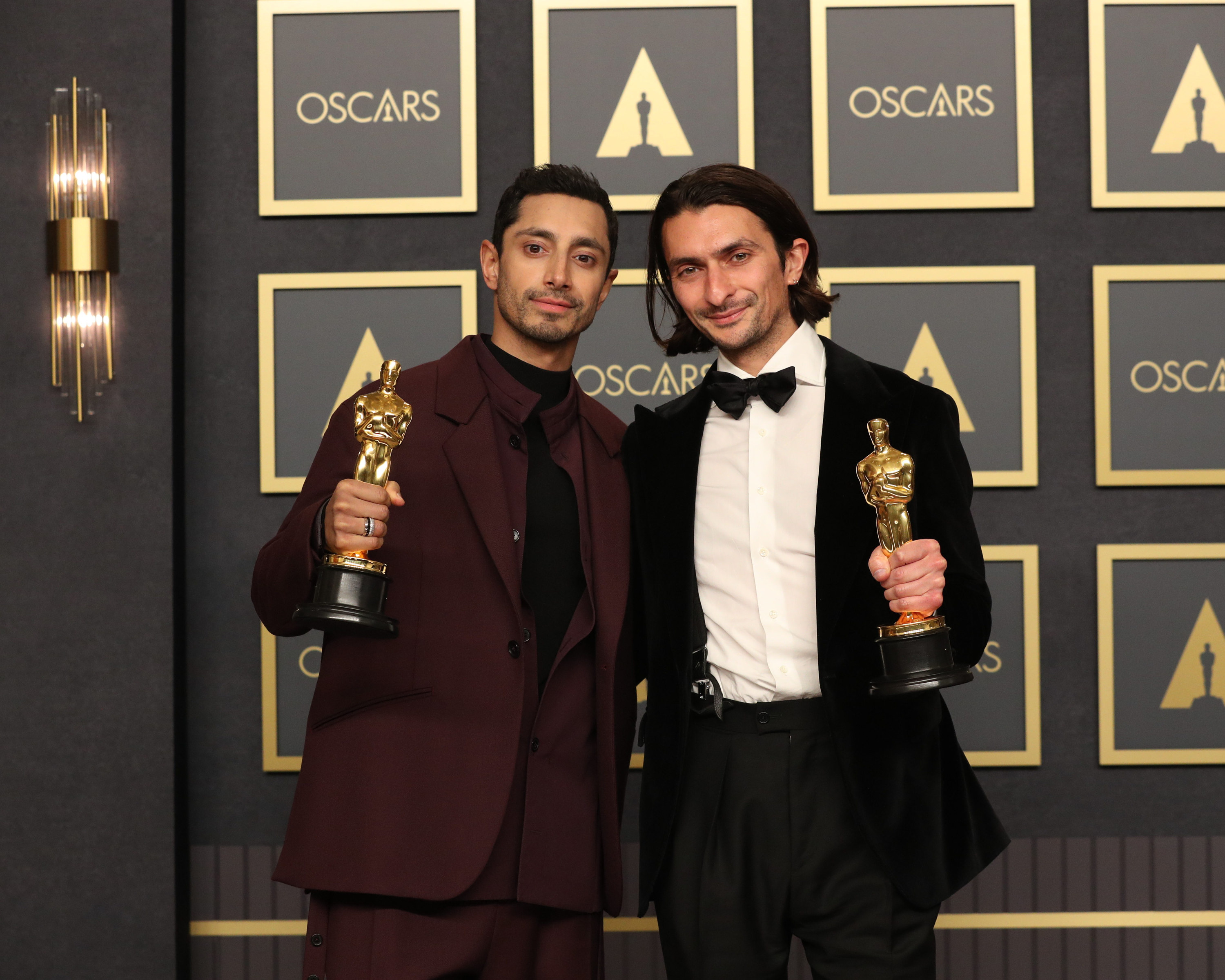 Riz holds his Oscar, with the wall behind him covered in black and gold Oscars posters