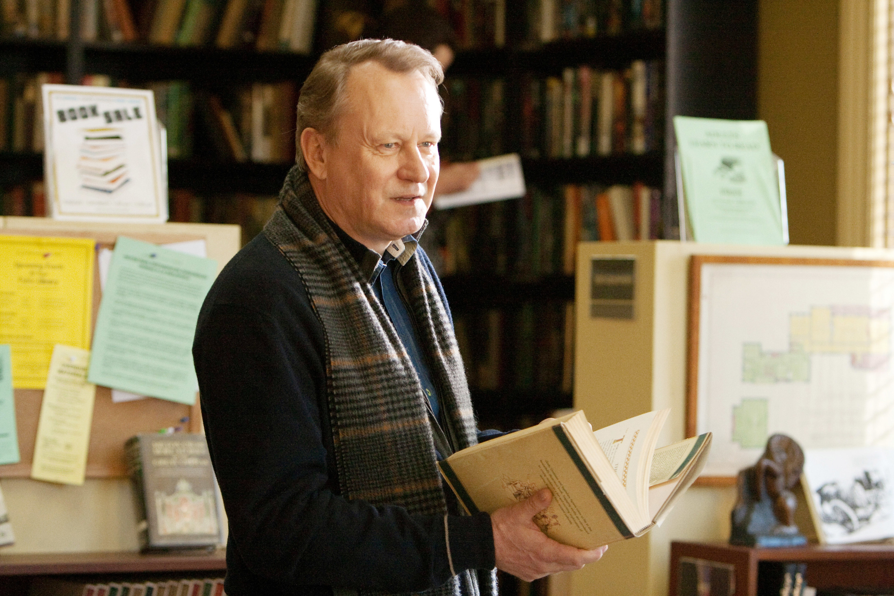 Stellan, wearing a scar, stands holding a book and with bookshelves behind him
