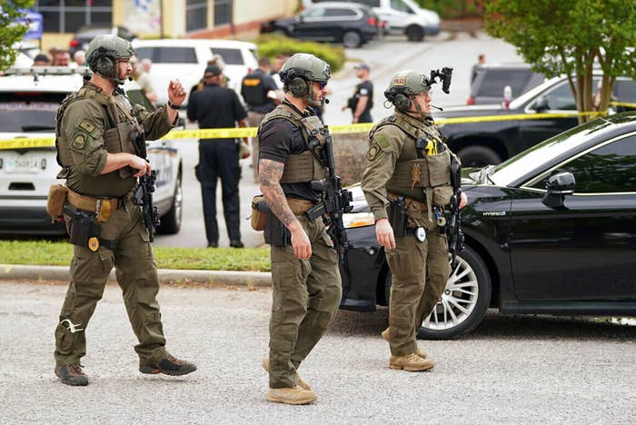 Three armed people in helmets and uniforms walk along the street with other officers and cars in the background