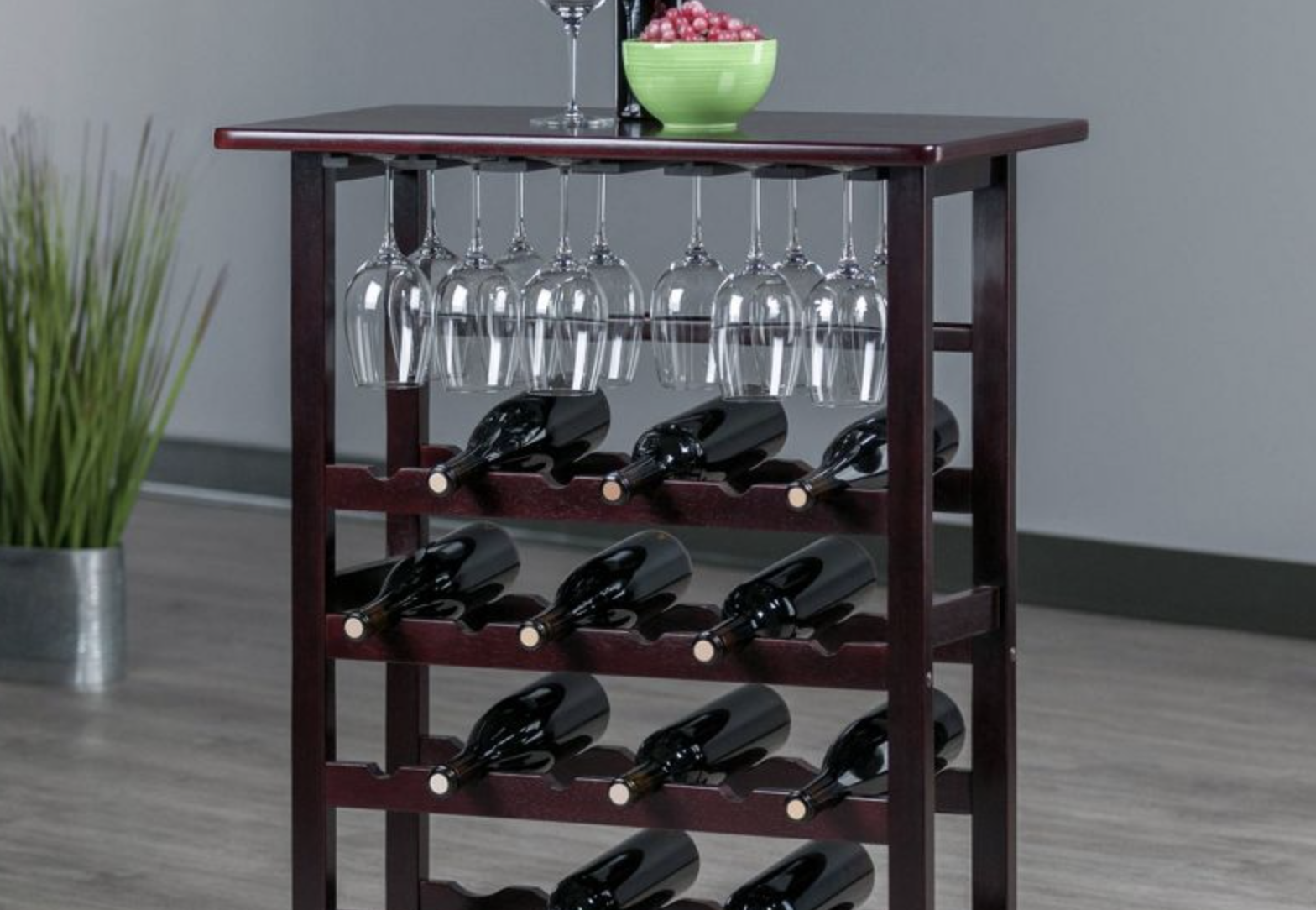 The wooden wine rack storing 11 bottles of wine vertically with wine glasses hanging from below tabletop. Wine bottle, wine glass and bowl of grapes served on top of rack