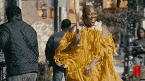 Titus Andromedon walks down the street in a brightly colored dress with fringe while swinging a baseball bat