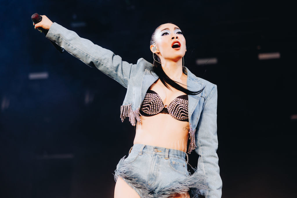 Rina Sawayama performing onstage at Coachella in a beaded bra, denim jacket, and denim shorts